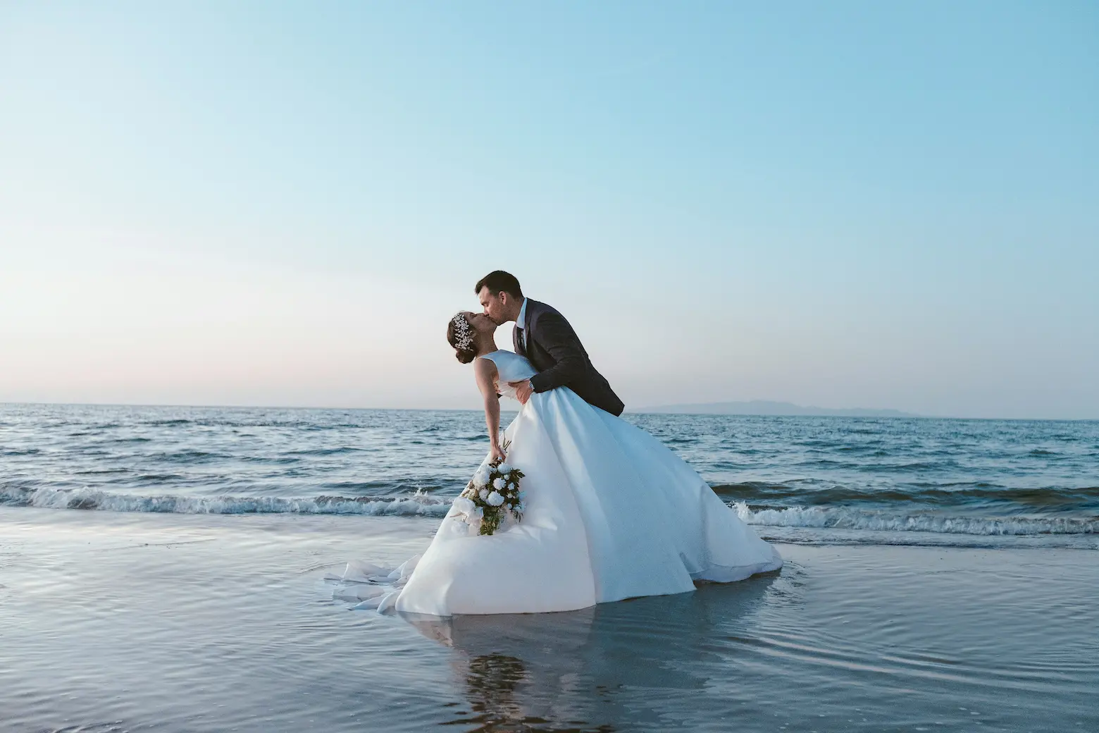 Deux jeunes mariés en tenue de mariage s'embrassent penchés, sur une plage les pieds dans l'eau de la mer.